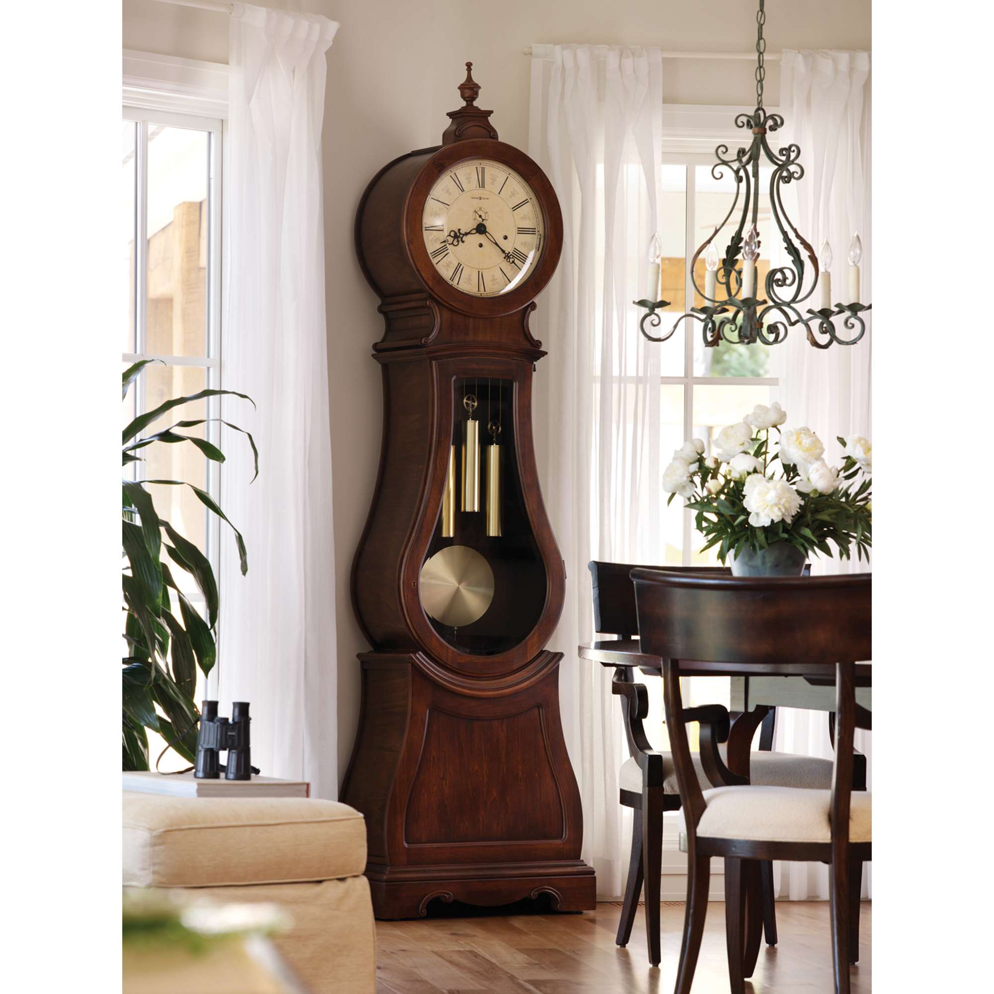 A tall, elegant grandfather clock in a bright dining area with a chandelier and dining table.
