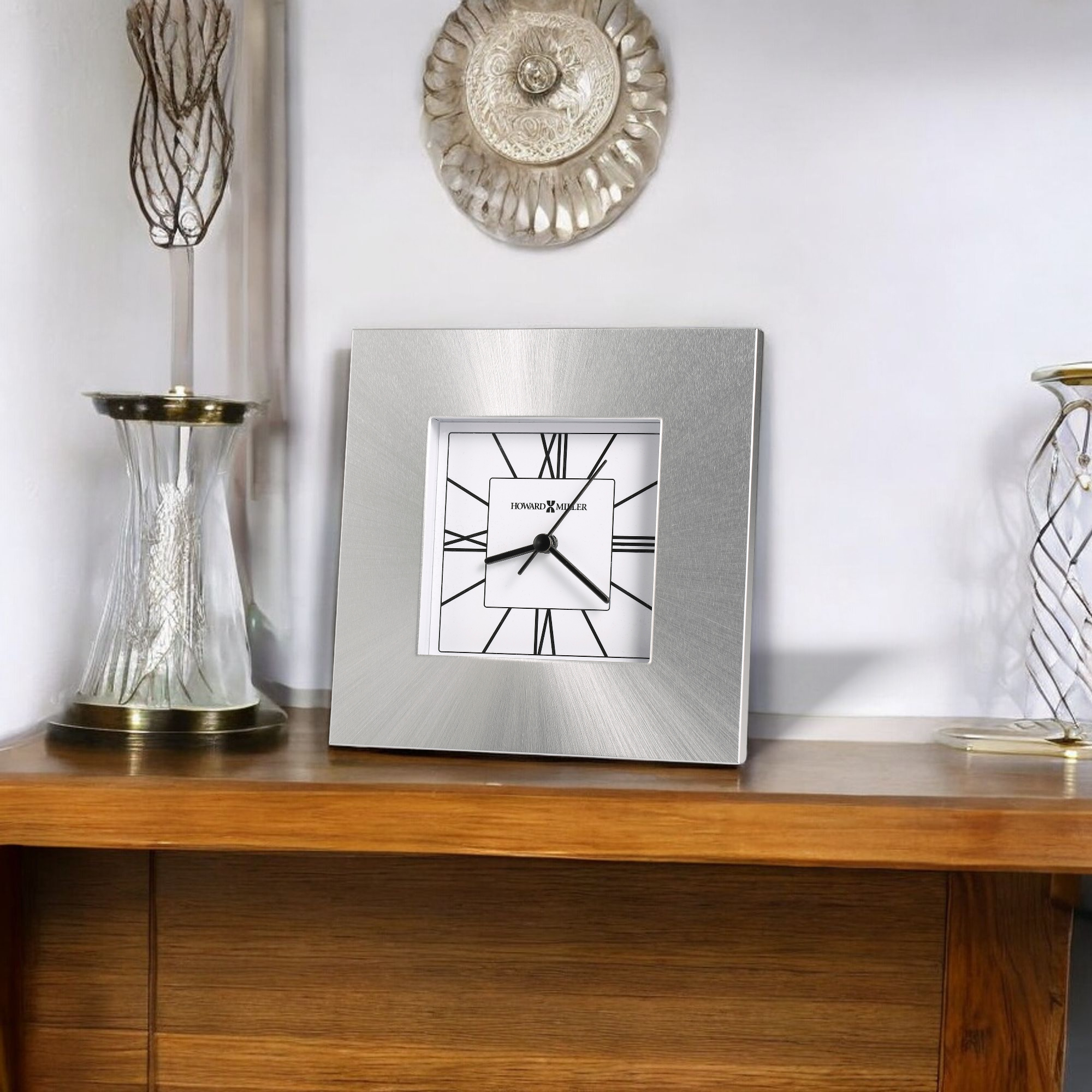 A modern square wall clock displayed on a wooden shelf with decorative vases.