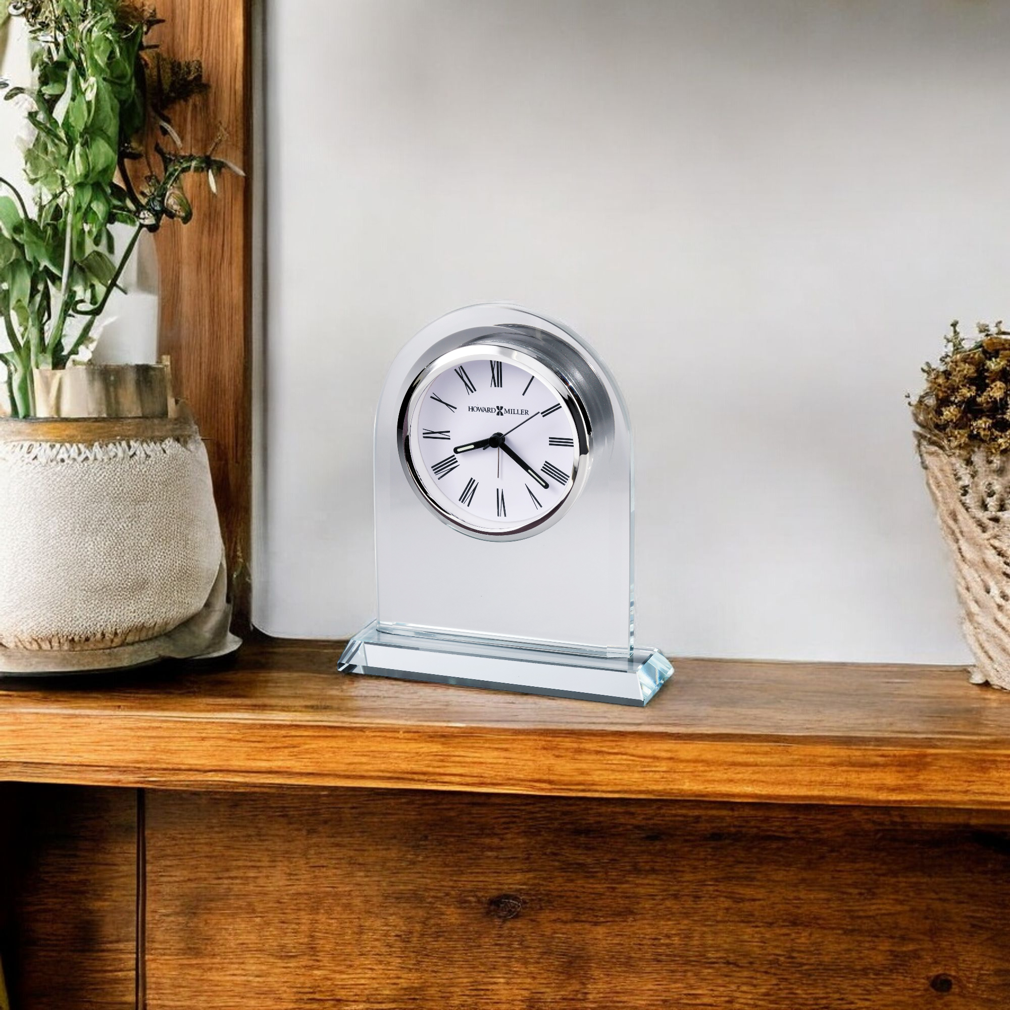 A sleek, modern desk clock with a clear case, displayed on a wooden shelf alongside a plant and decorative items.