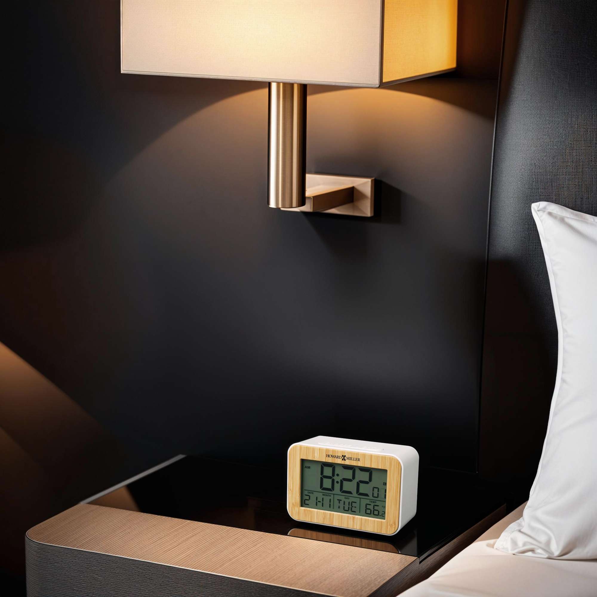 A digital alarm clock displaying the time on a nightstand next to a lamp.