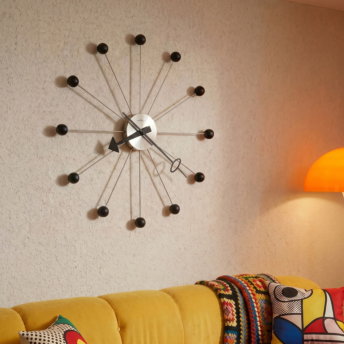 Modern wall clock on a beige wall with a yellow sofa and colorful cushions in the foreground.