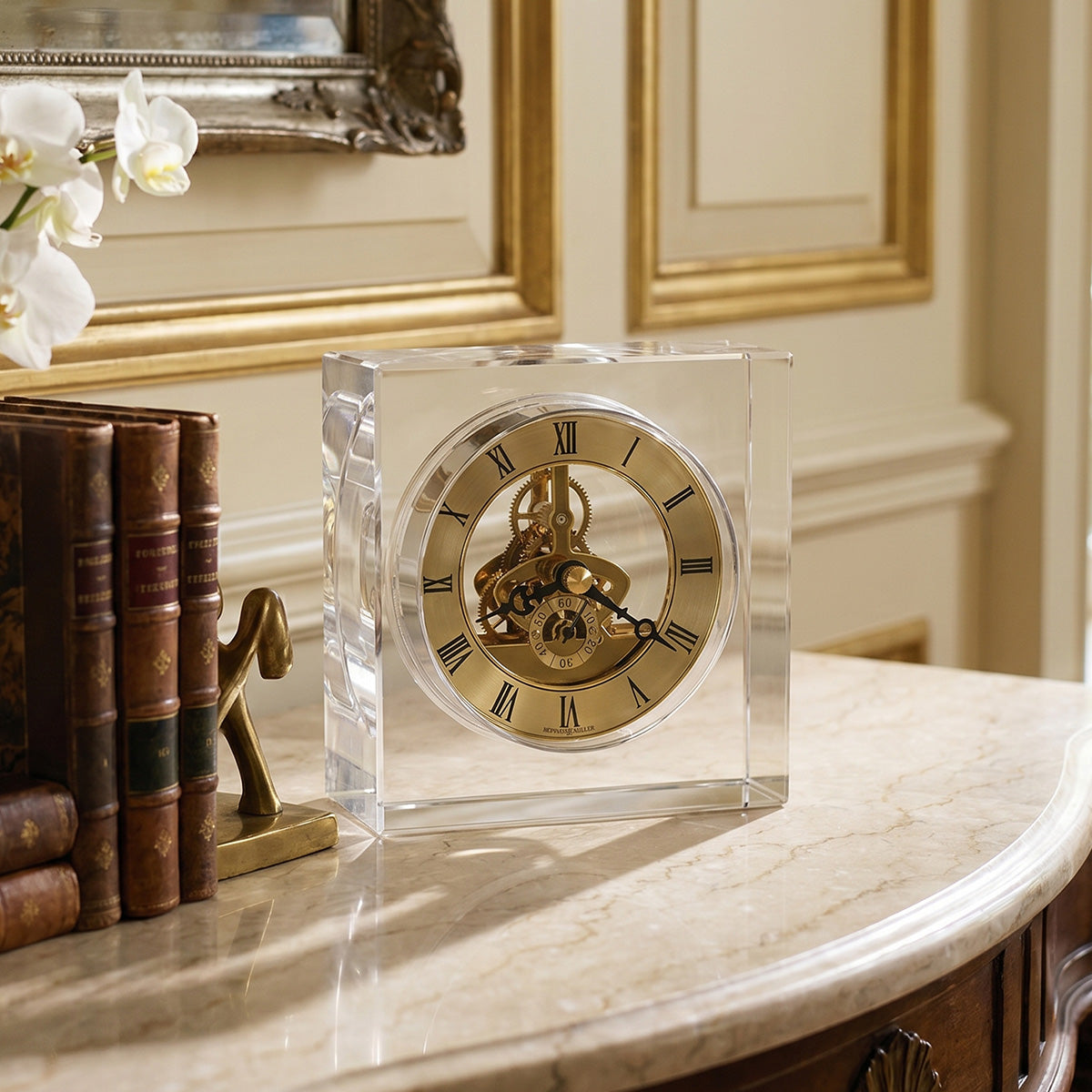 Decorative clock with glass casing on a marble surface, surrounded by books and flowers.