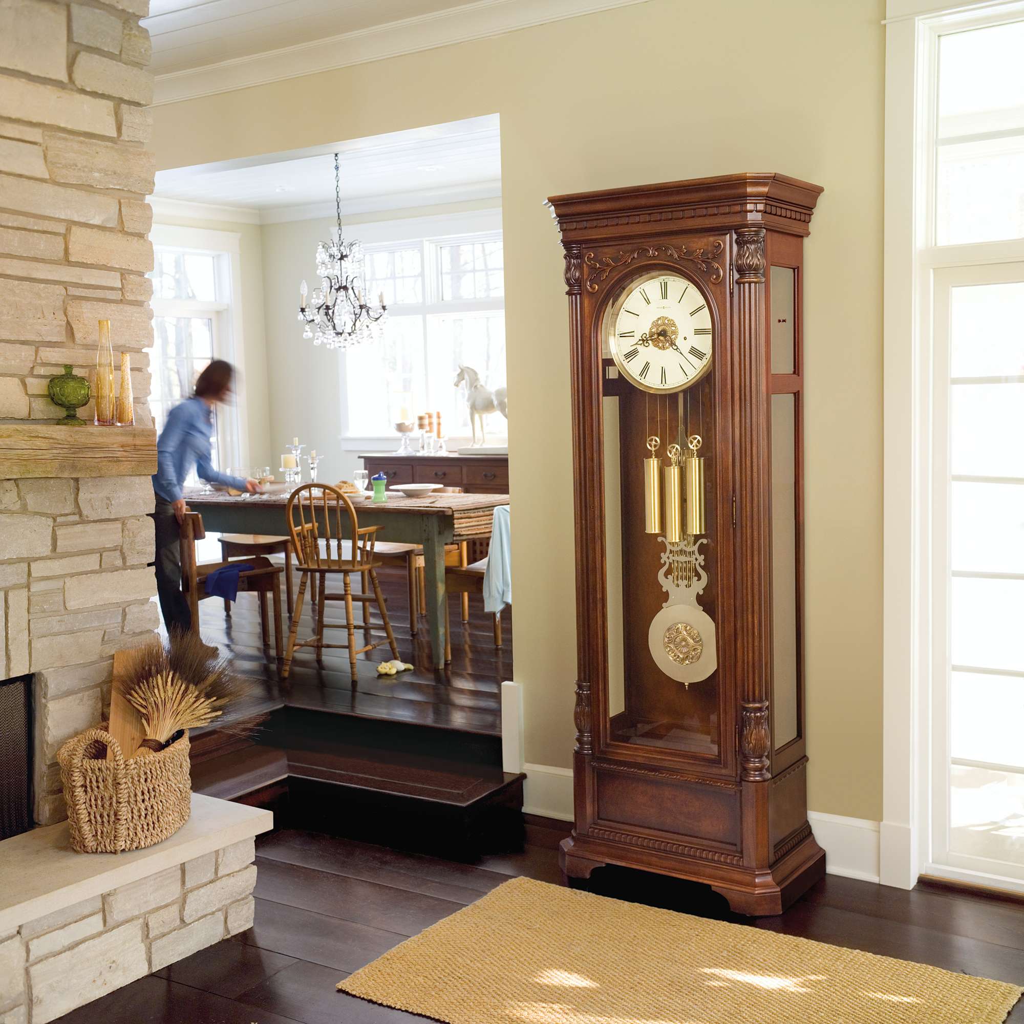 A wooden grandfather clock stands prominently in a room with a dining area in the background.