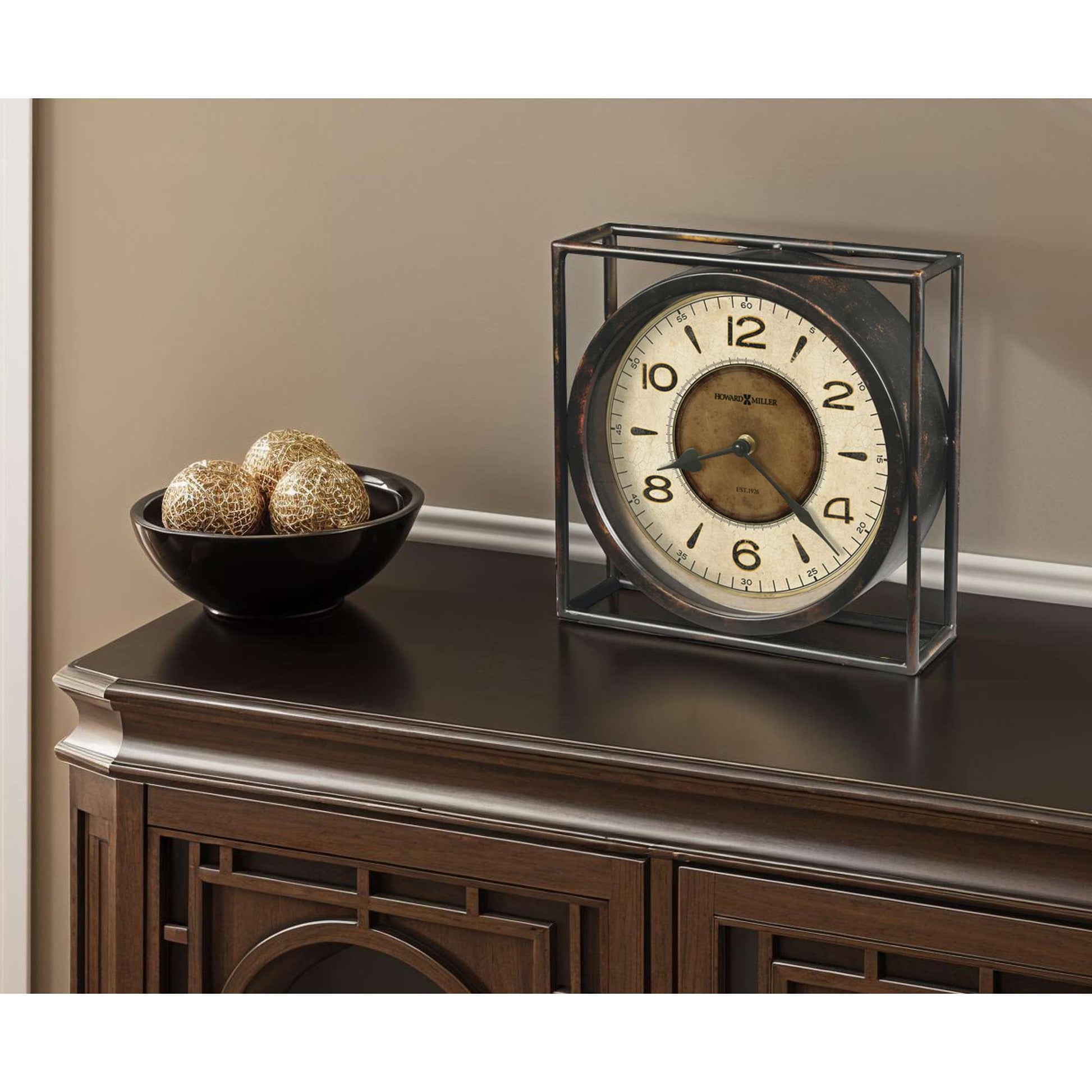 A vintage-style clock in a glass frame on a dark surface beside a bowl of decorative orbs.