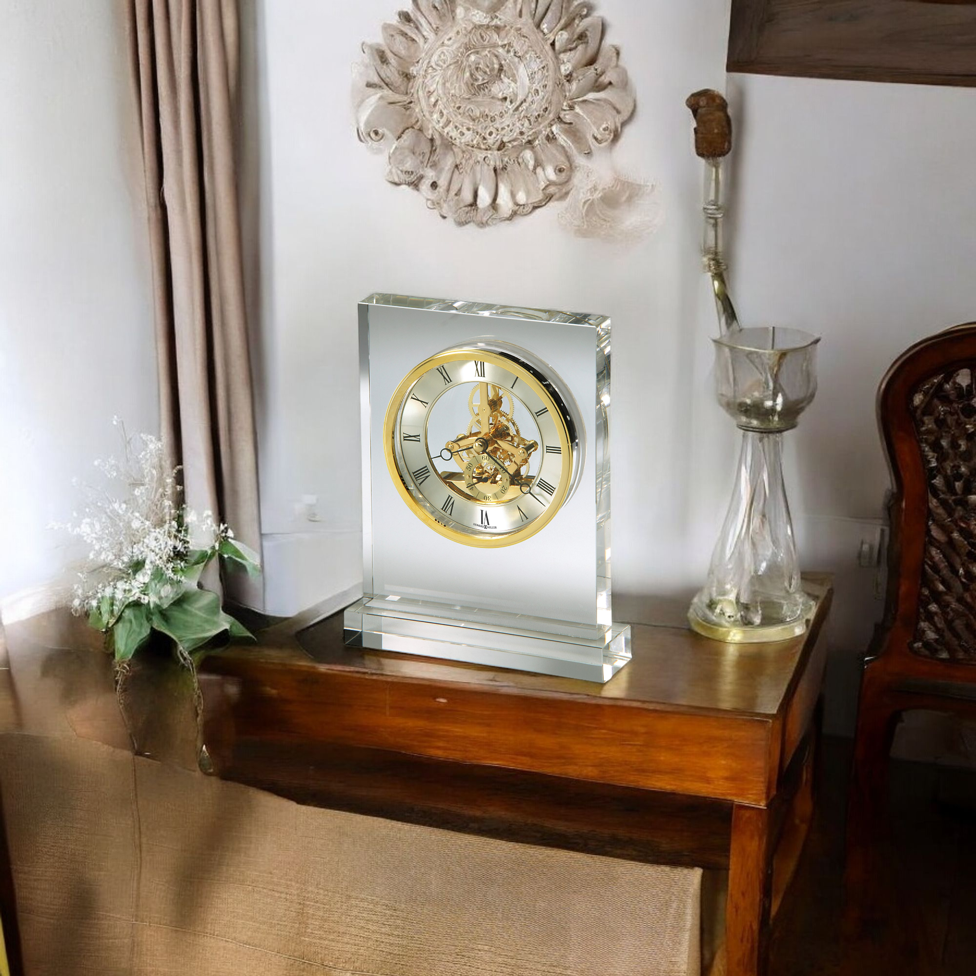 A decorative clock on a wooden table with a floral arrangement and vintage glassware.