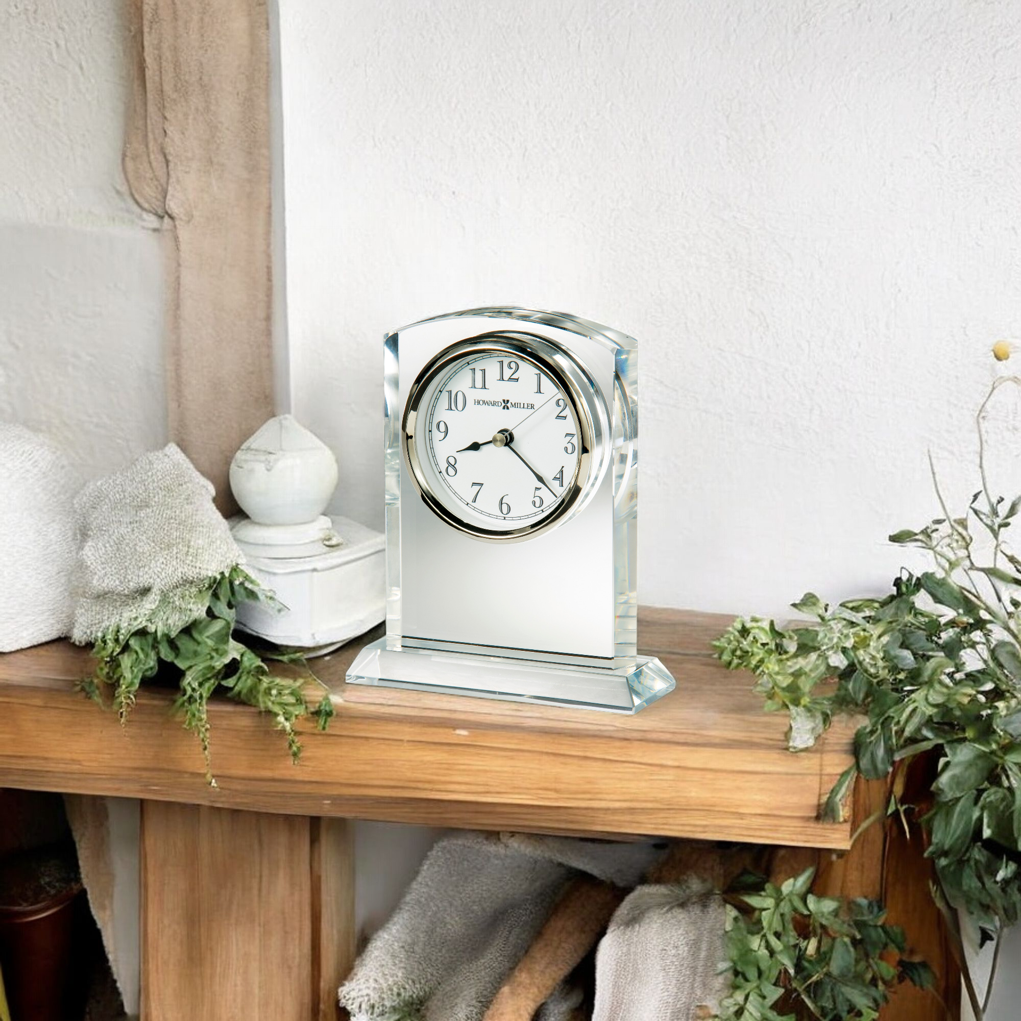 A decorative clock on a wooden shelf with plants and a white towel.