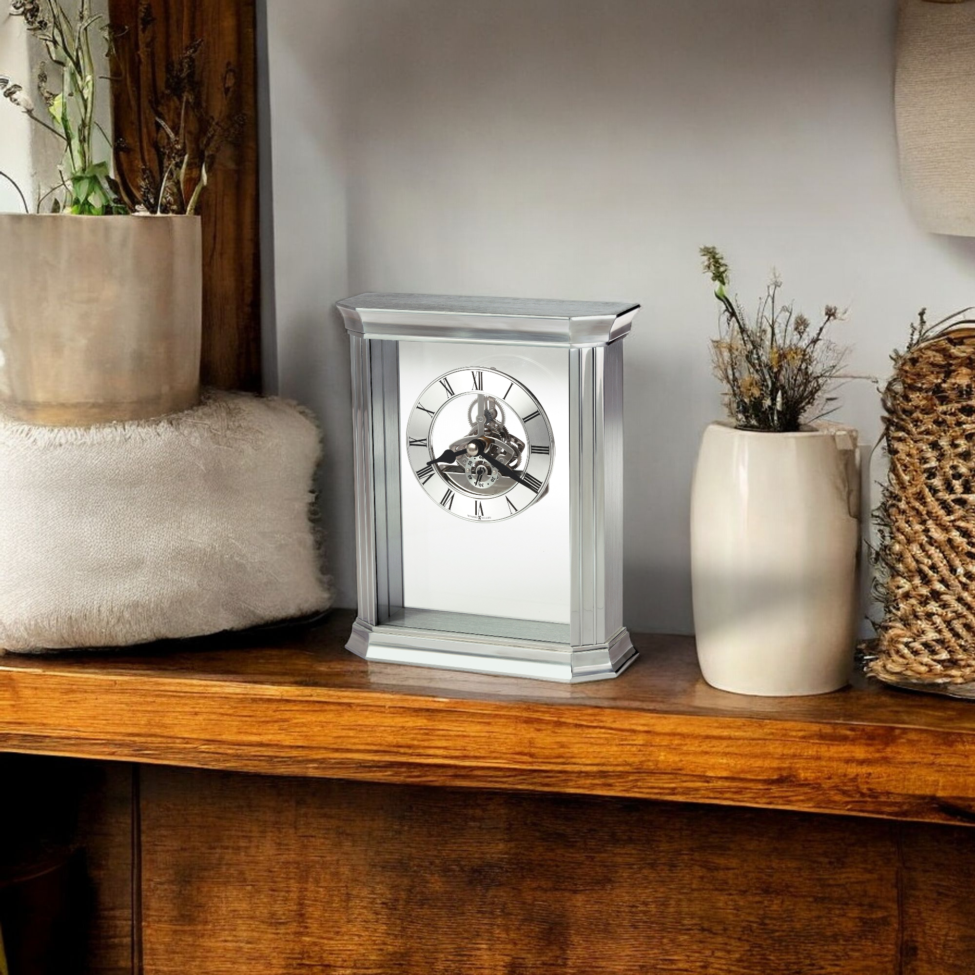 A decorative clock on a wooden shelf, surrounded by plants and a woven basket.