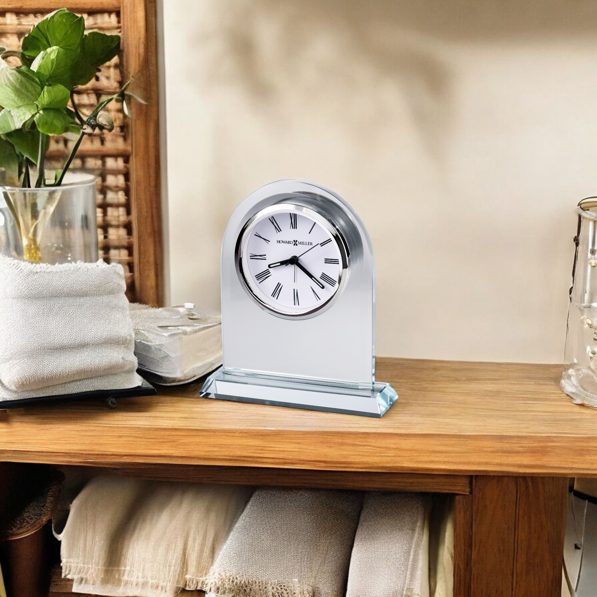 A decorative clock placed on a wooden shelf with a plant and folded towels.