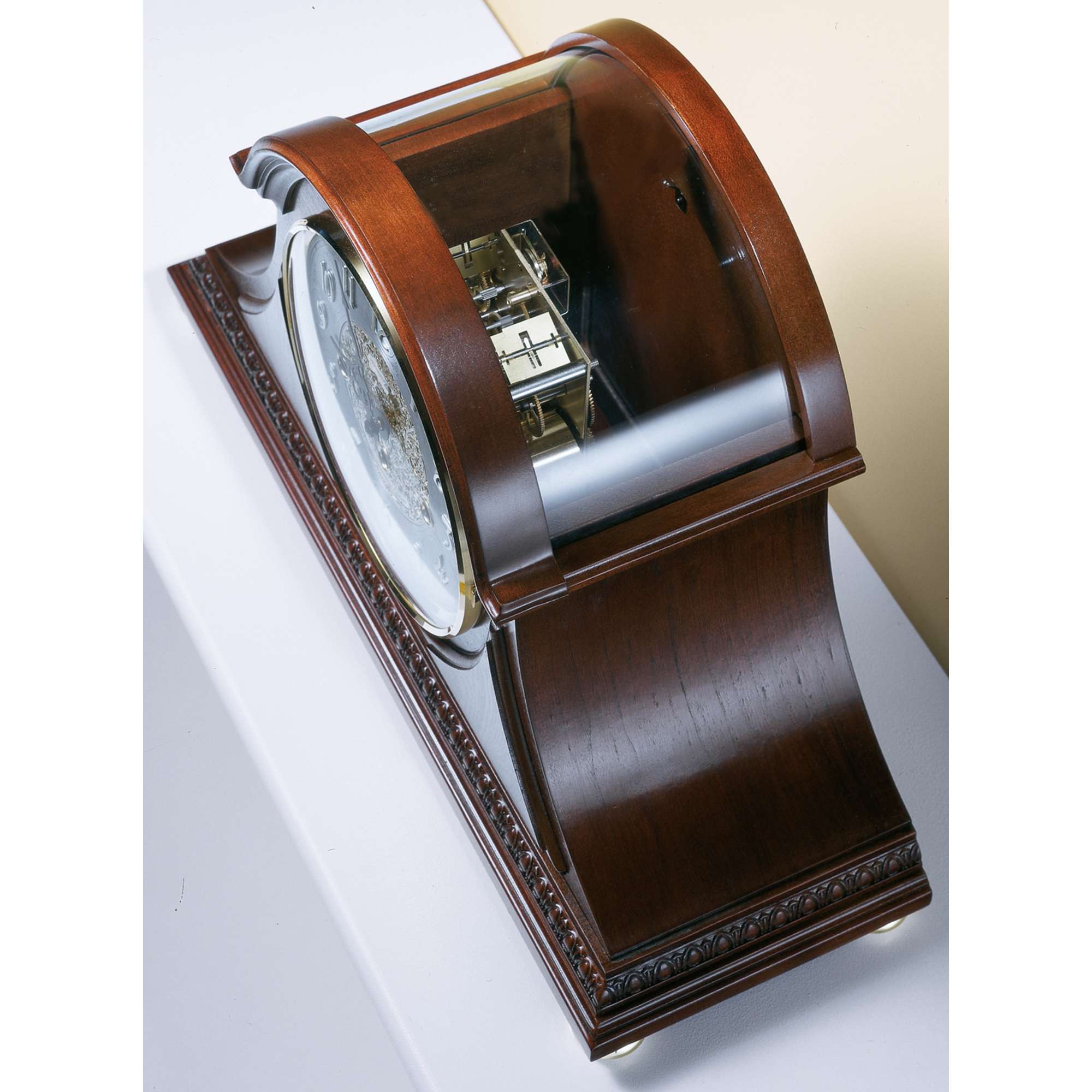 A wooden mantel clock with a curved design, displaying intricate mechanisms behind a glass cover.